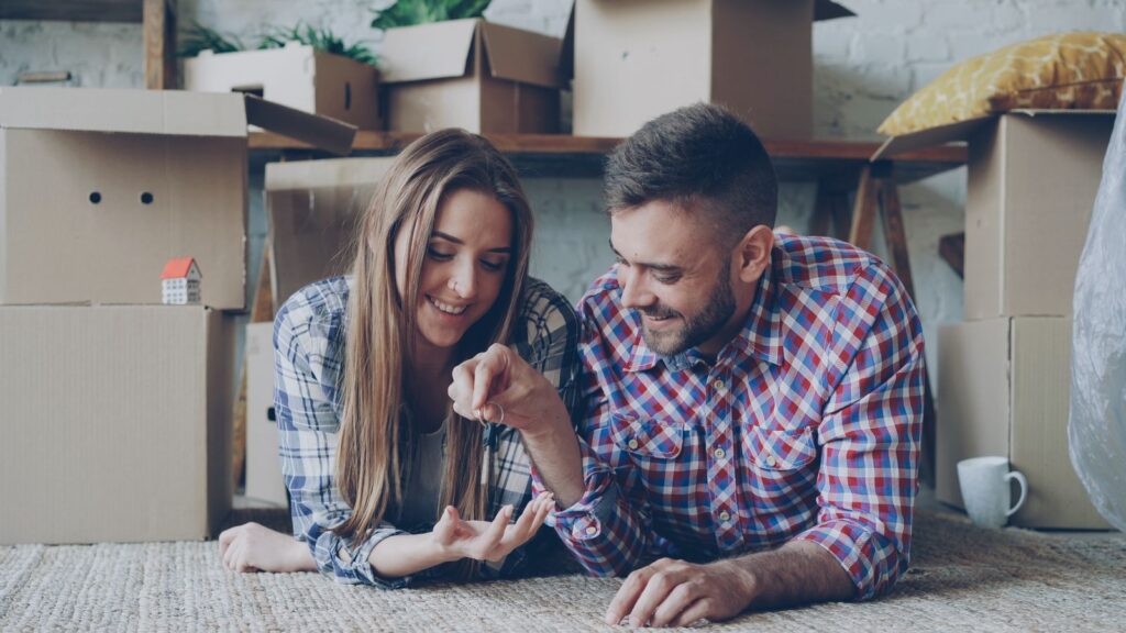 A young couple sitting among moving boxes in their first home, holding the keys.