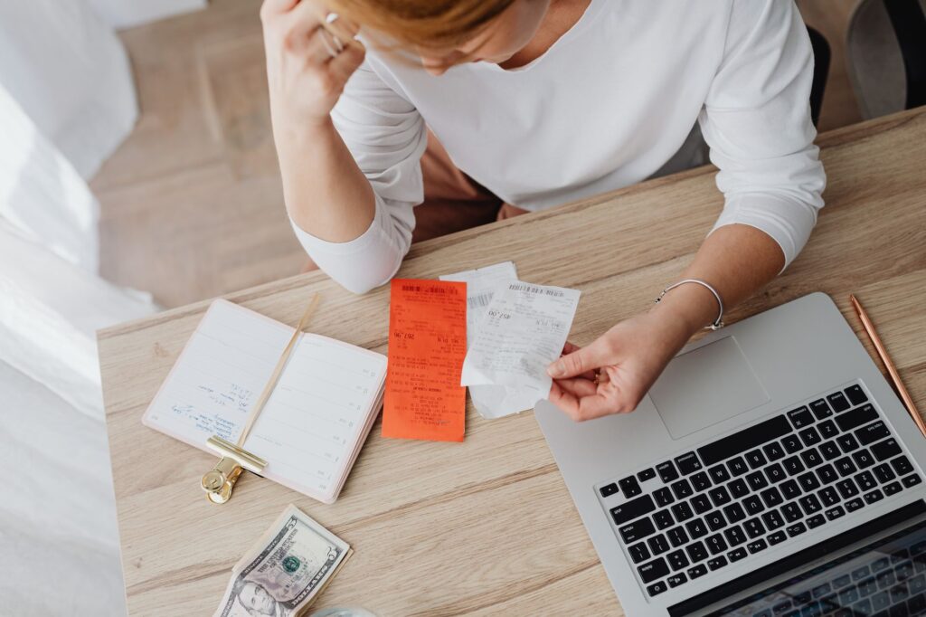 A woman sitting at a kitchen table checking her savings account balance on a laptop while holding a bank statement.