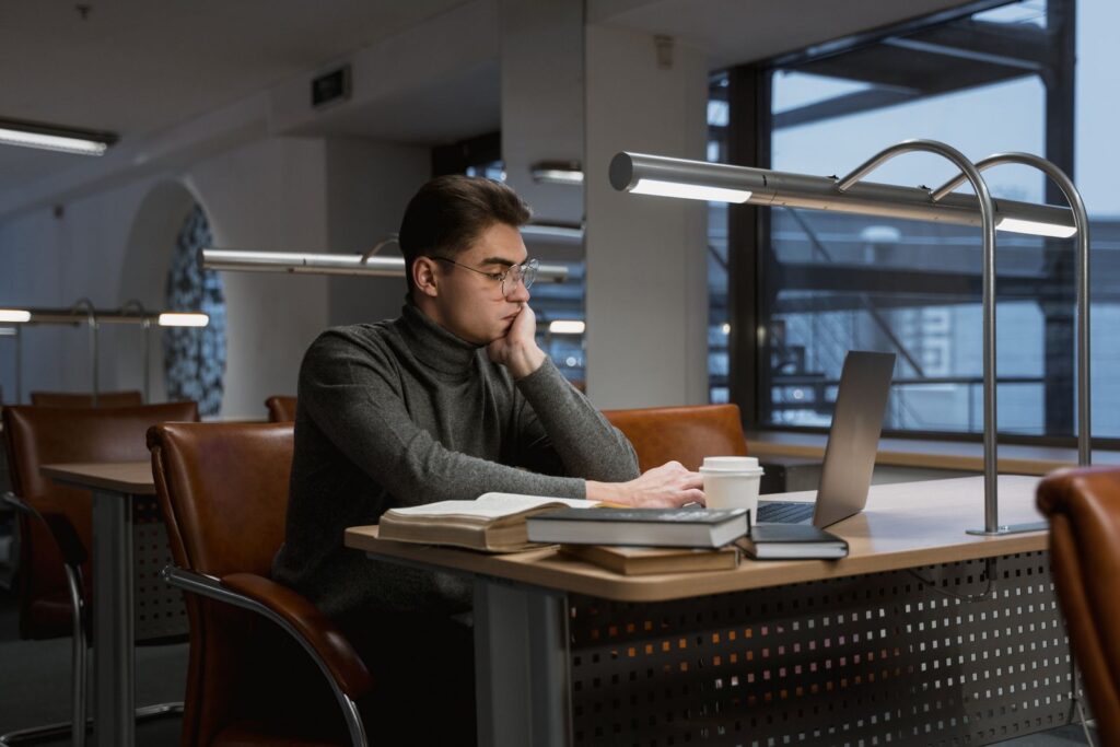 A university student working at a laptop with books and notes spread across the desk.
