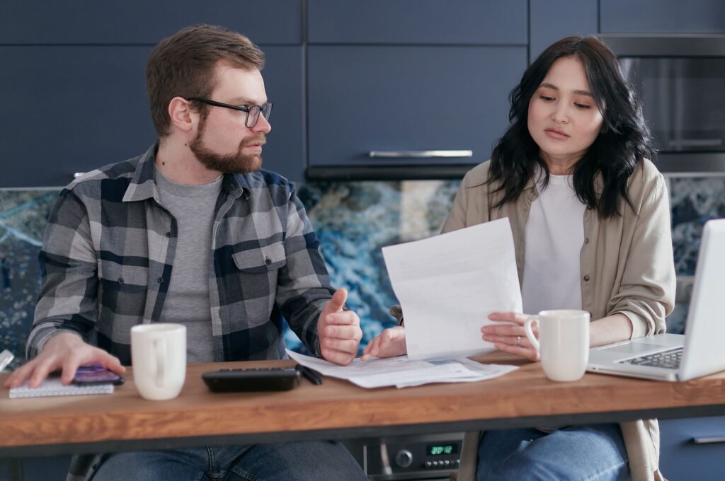 A parent at a kitchen table with a laptop and household paperwork, working out a monthly benefits and bills budget.