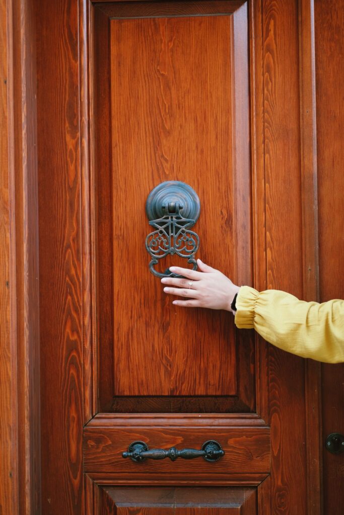 A person in a yellow sleeve reaching towards an ornate metal door knocker on a wooden front door.