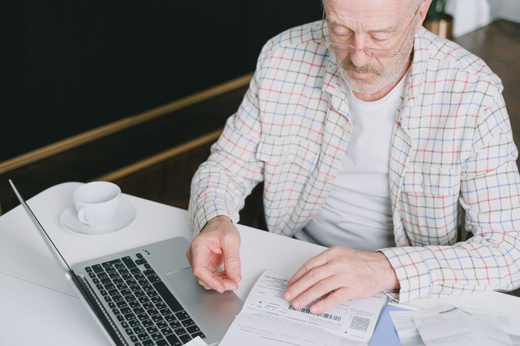 An older man in glasses reading a document at a desk beside an open laptop.