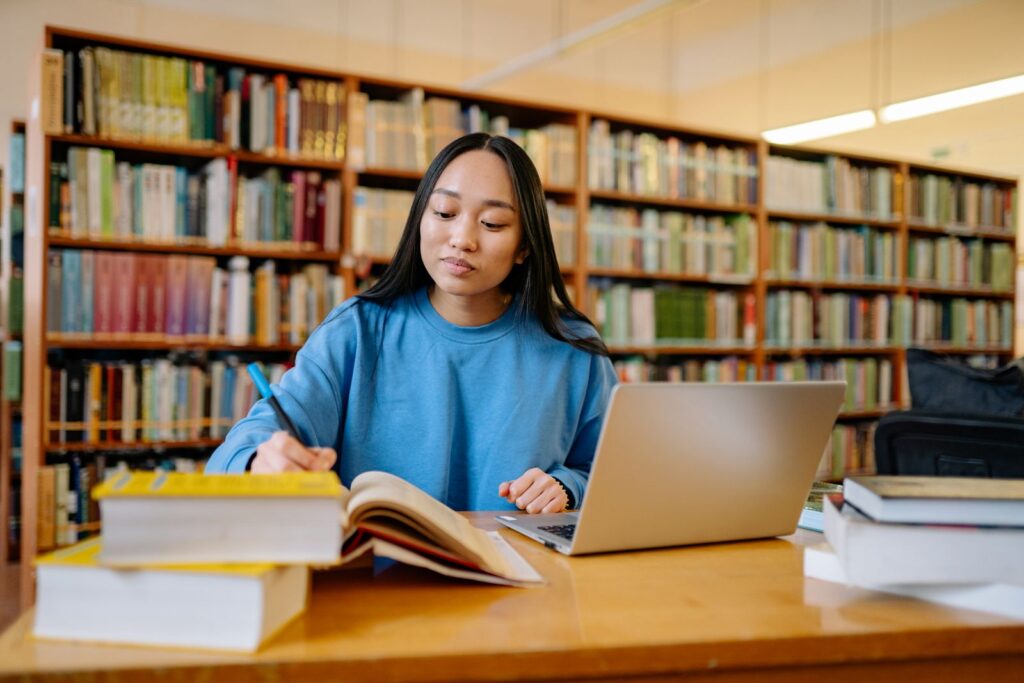 A young woman sitting at a library desk, working on a laptop with open textbooks beside her.