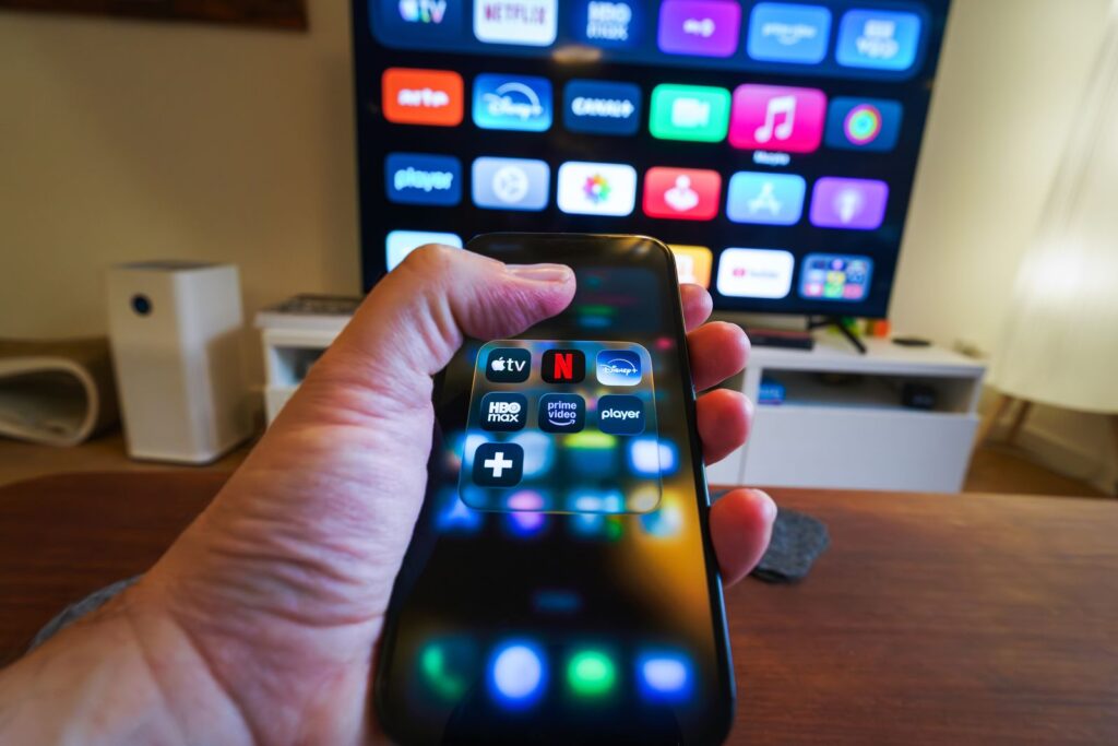 A hand holding a smartphone displaying streaming app icons in front of a television screen showing apps.